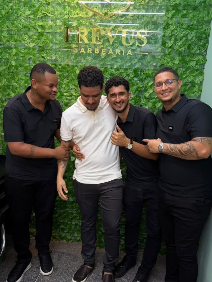 Kelson, Júnior e Hariel abraçados na inauguração, em frente ao letreiro dourado da Trevus na parede de folhagem.