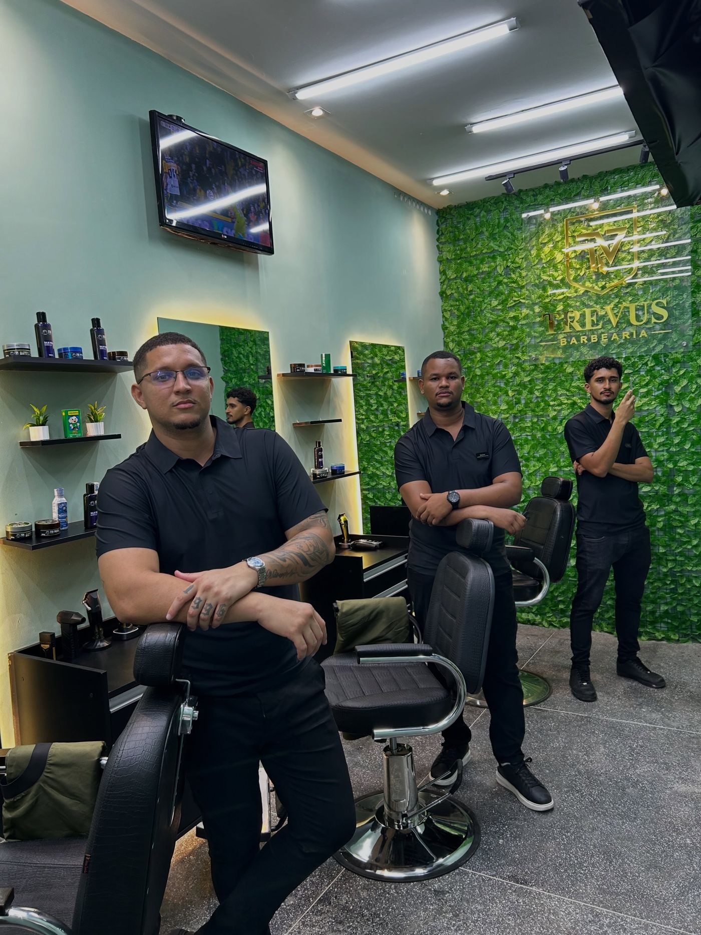 Hariel, Júnior e Kelson no salão reformado da Trevus, entre as cadeiras de couro e a parede de folhagem com o letreiro dourado.